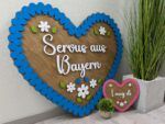 Decorative wooden heart-shaped sign with a bright blue scalloped border, white flowers and green leaves, and cursive white lettering, mounted against a wall beside plants.