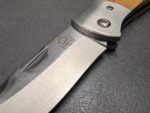 Close-up of a stainless steel knife blade with a wooden handle, showing screws and the PUMATEC logo on the blade edge area.