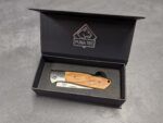 Black presentation box open to reveal a folding knife with a wooden handle; Puma TEC branding on the inner lid.